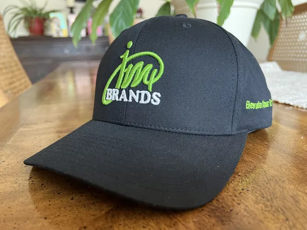 Black cap with the green JM4 Brands logo embroidered on the front, on a wood table