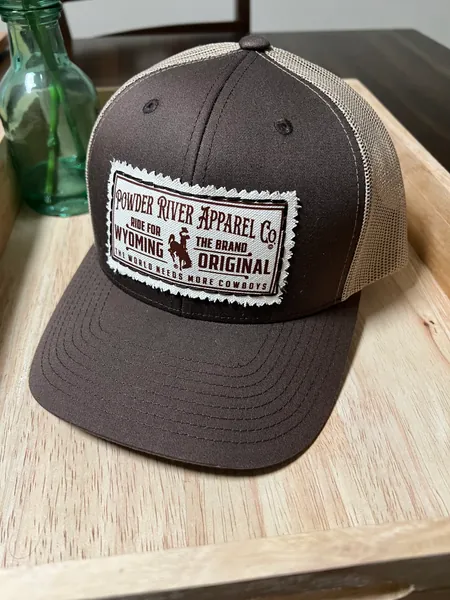 Brown and khaki trucker cap with a Powder River Apparel Co. patch showing a bucking horse and rider